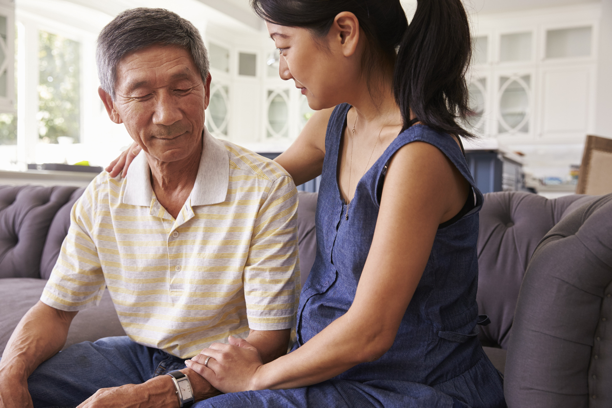 Father and daughter sitting on the couch together, with one of the daughter’s arms over her father’s shoulder and the other touching his forearm.  